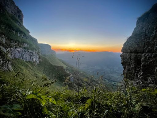 Paisaje montañoso al amanecer, con sol saliendo entre las montañas y vegetación.